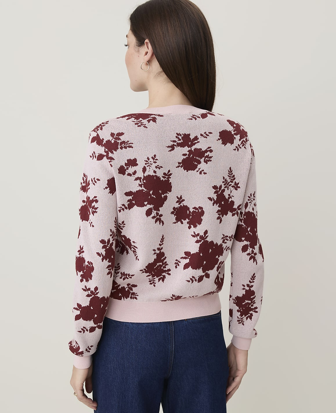 A woman in a pink floral sweater and blue jeans stands against a plain, light background.