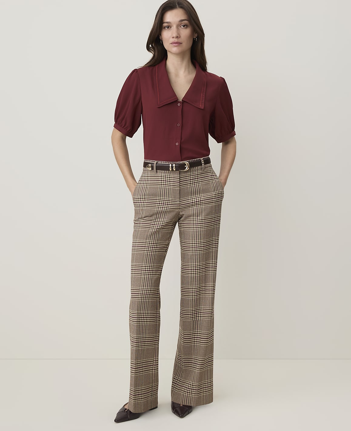 A woman in a maroon blouse and plaid pants stands against a plain, light-colored background.