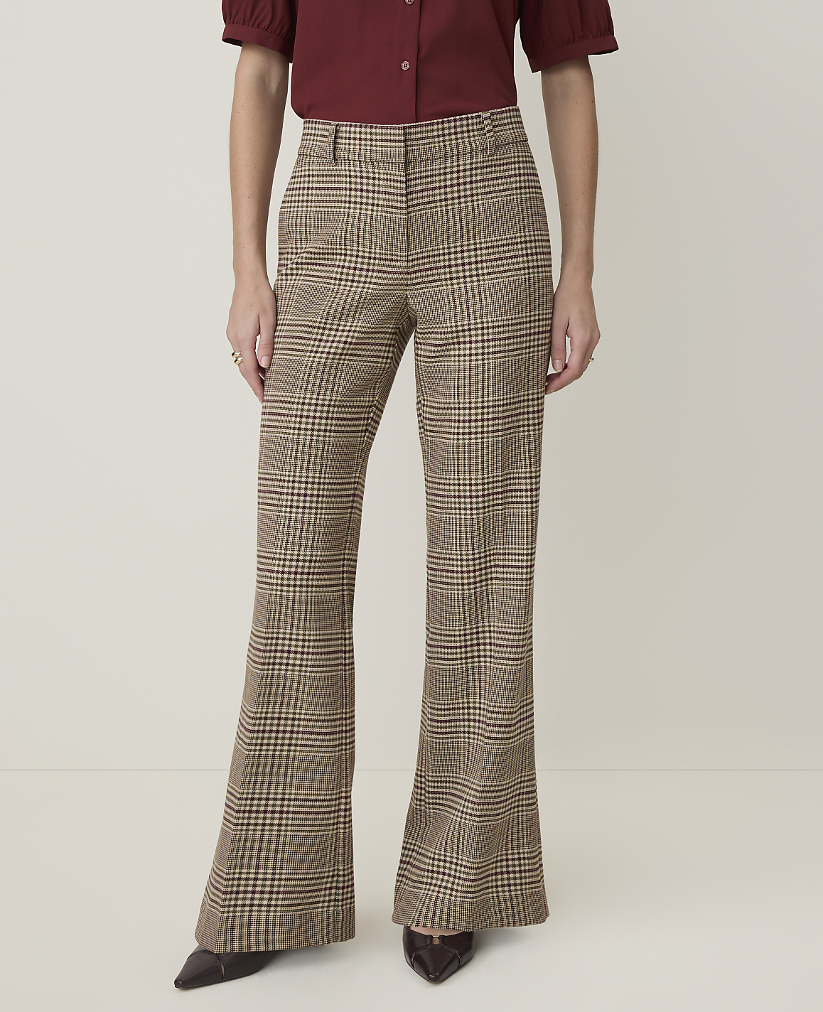 A person stands in flared plaid trousers and a maroon blouse against a plain, light-colored background.