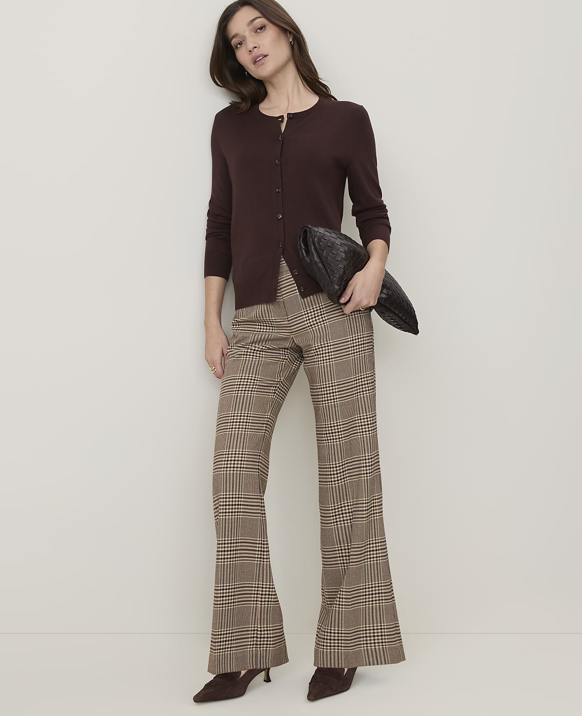 A woman in a brown cardigan and plaid flared pants stands against a plain, light-colored wall, holding a dark clutch.