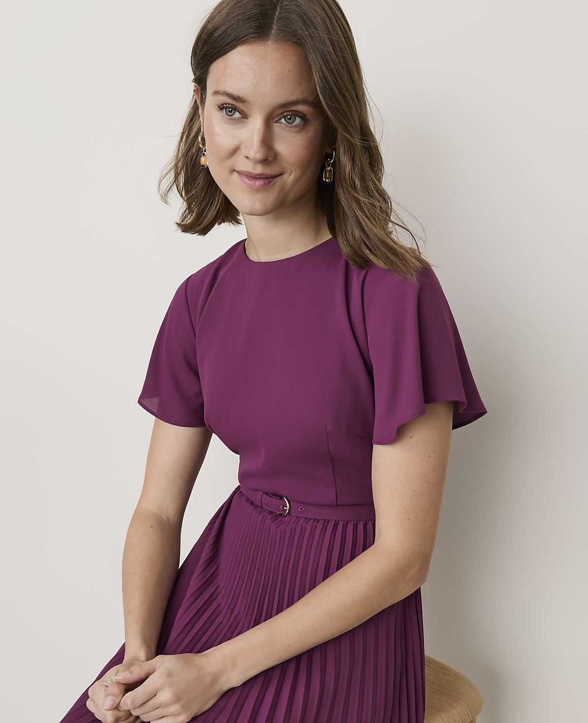 A woman in a purple dress with flutter sleeves and a pleated skirt sits against a plain, light-colored background.