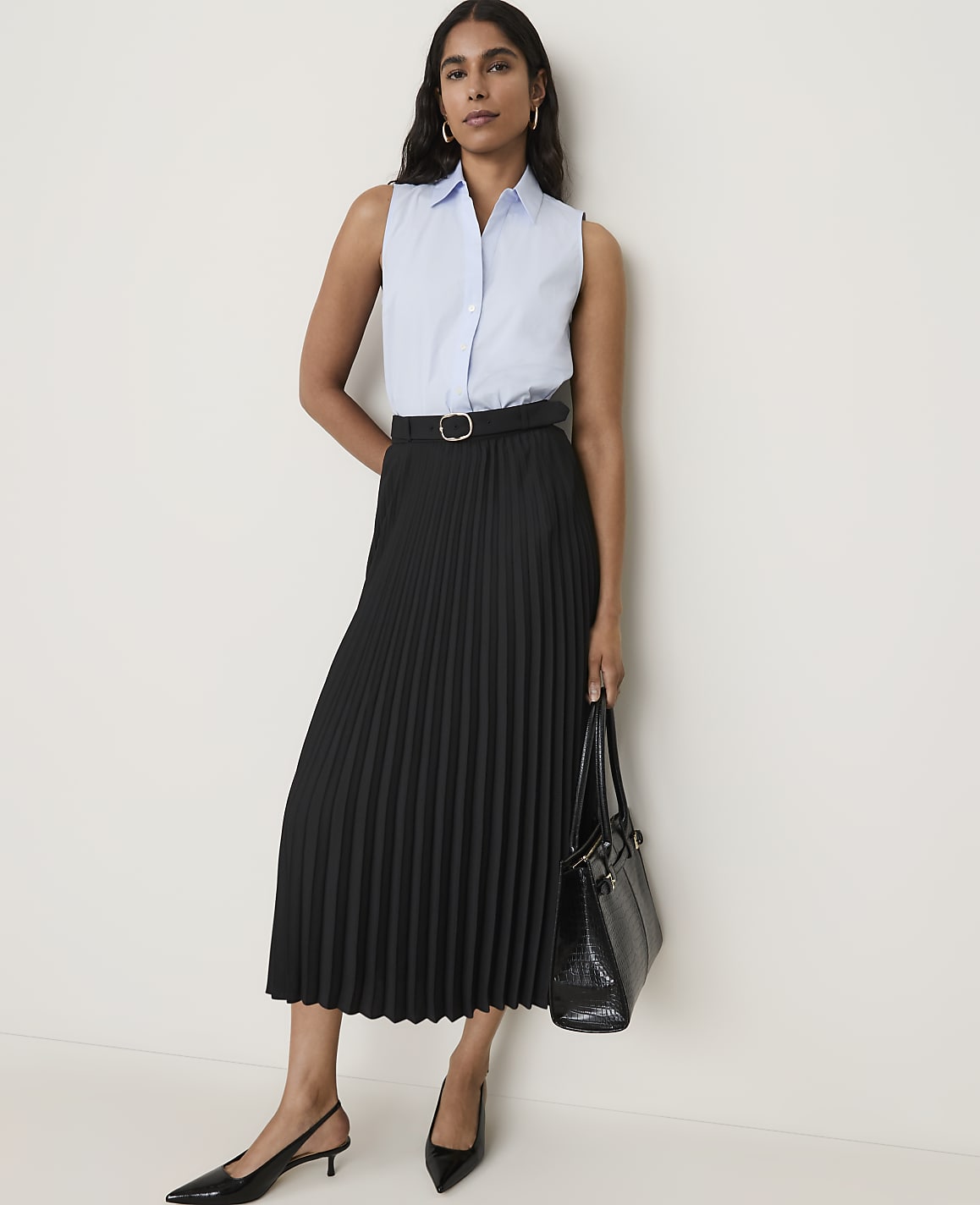 A woman in a light blue sleeveless shirt and a black pleated skirt stands against a plain white background, holding a black handbag.