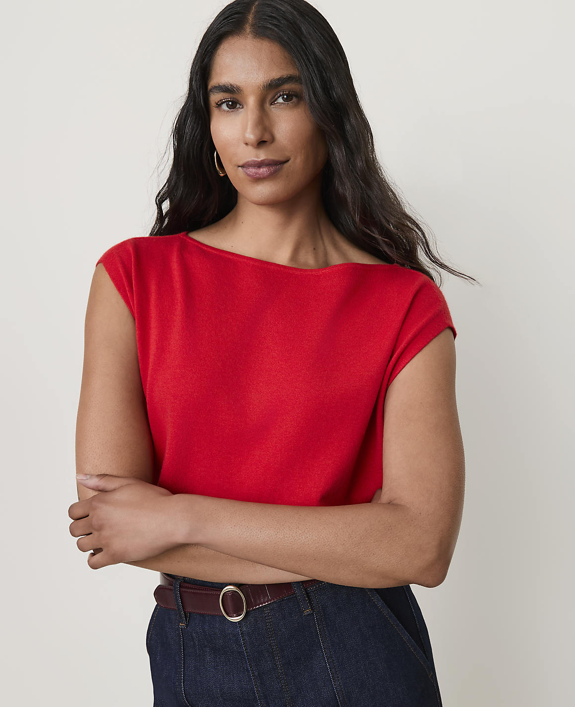 A woman in a red top and dark jeans stands against a plain background.