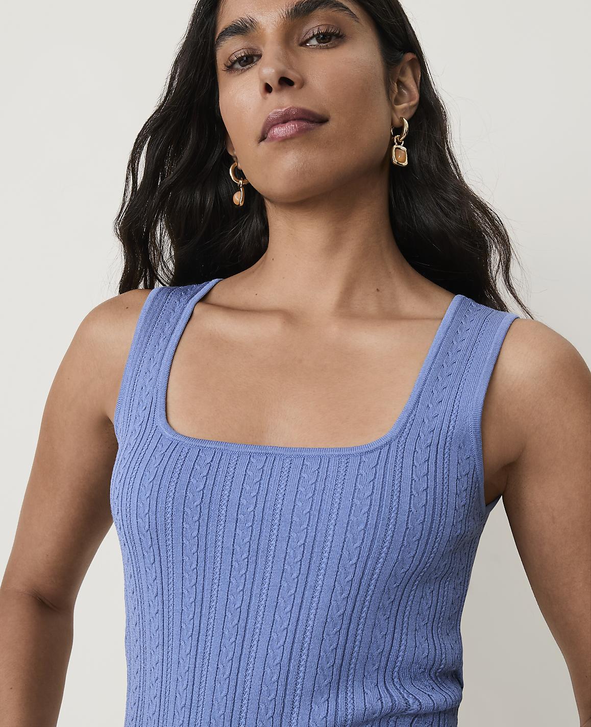 A woman with dark hair and earrings wears a blue cable-knit tank top against a plain background.