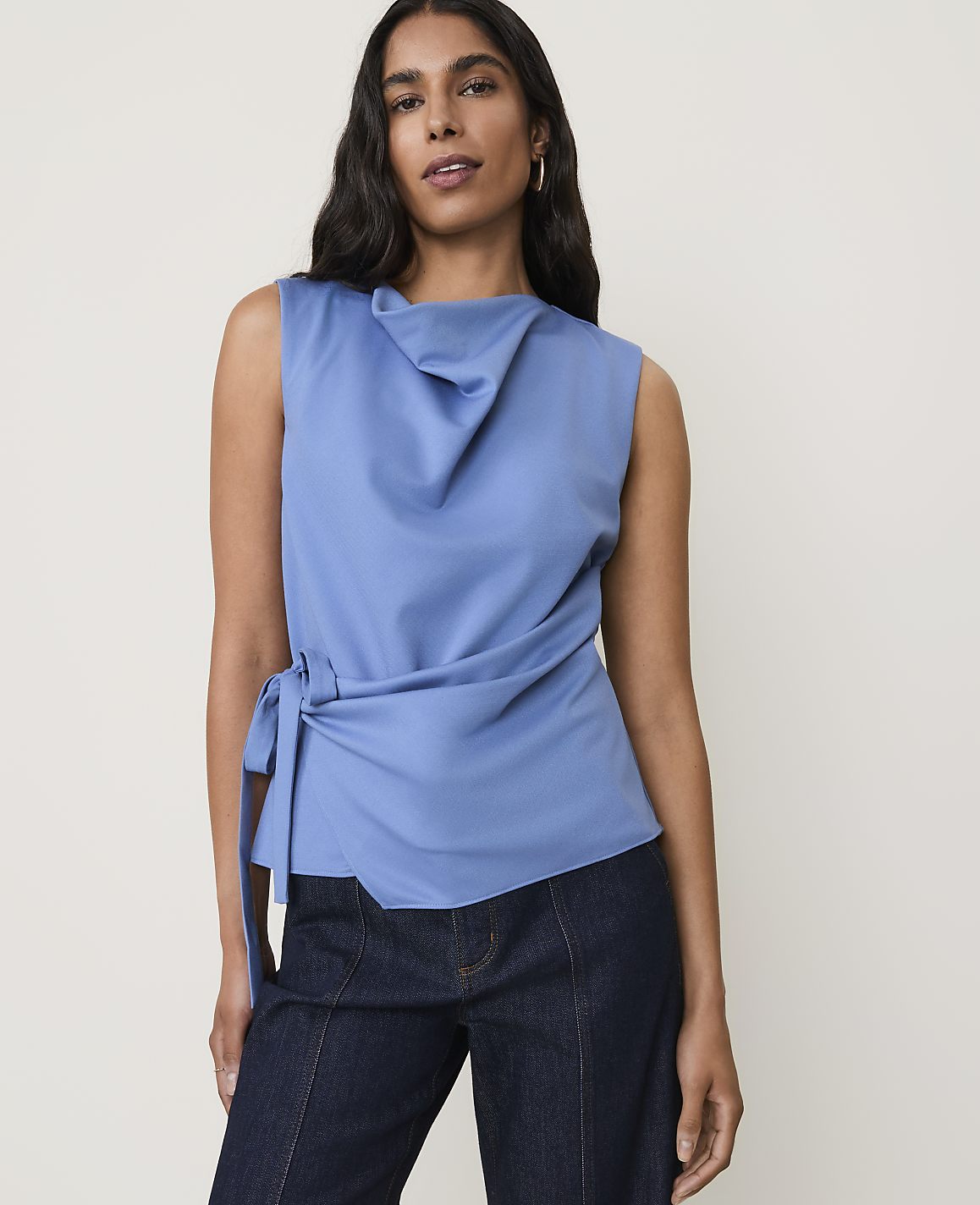 A woman in a blue cowl-neck top and dark jeans stands against a plain, light-colored background.