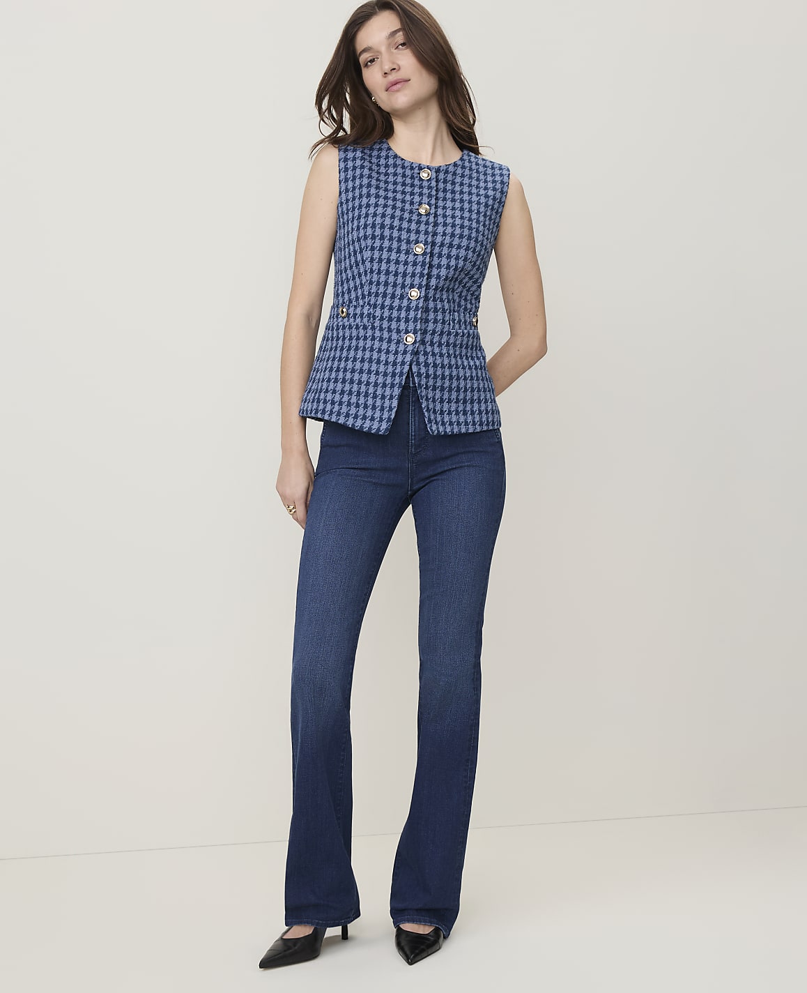 A woman in a blue tweed vest and dark wash flared jeans stands against a plain, light-colored background.