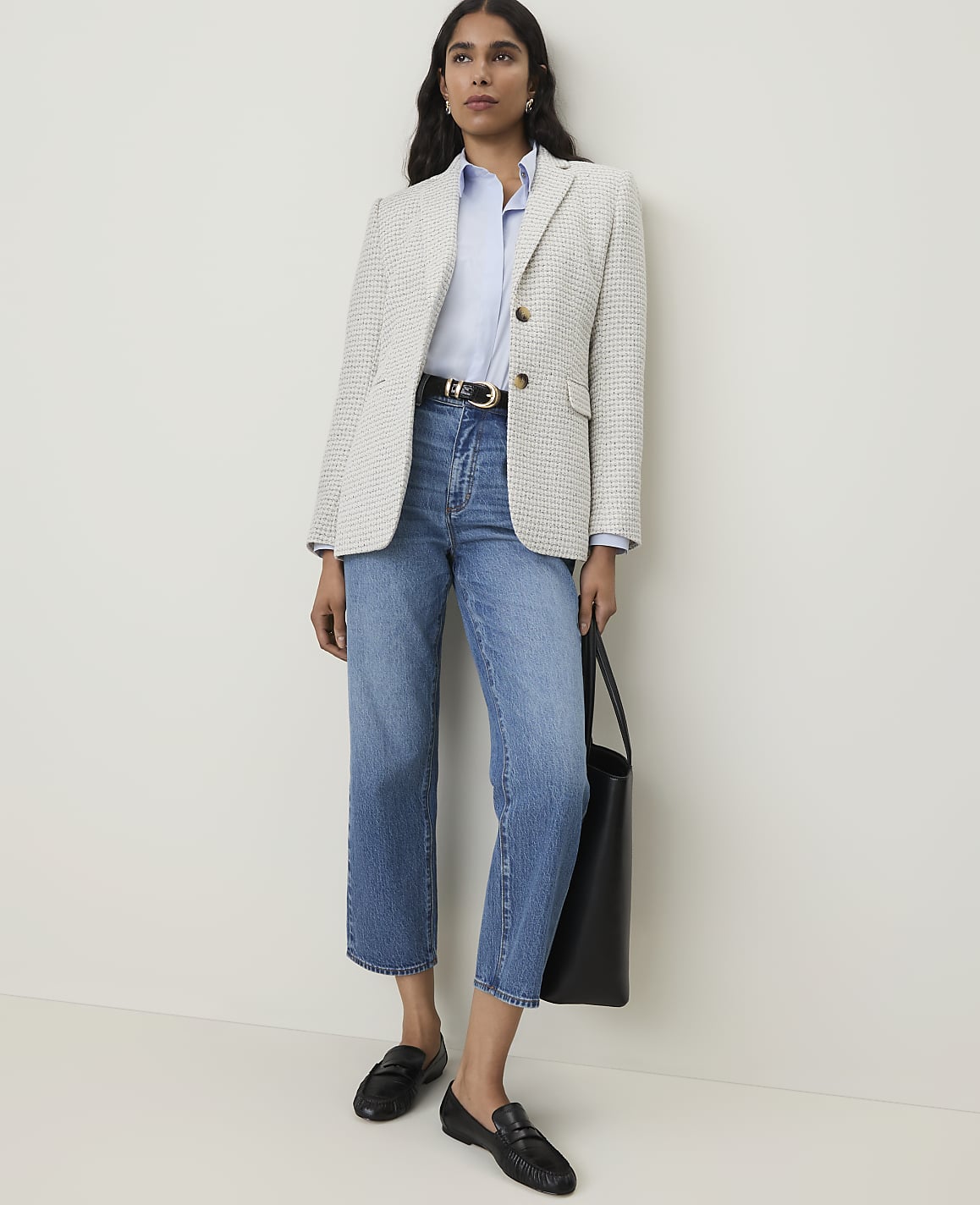 A woman in a light-colored tweed blazer, blue shirt, and jeans stands against a plain white wall.
