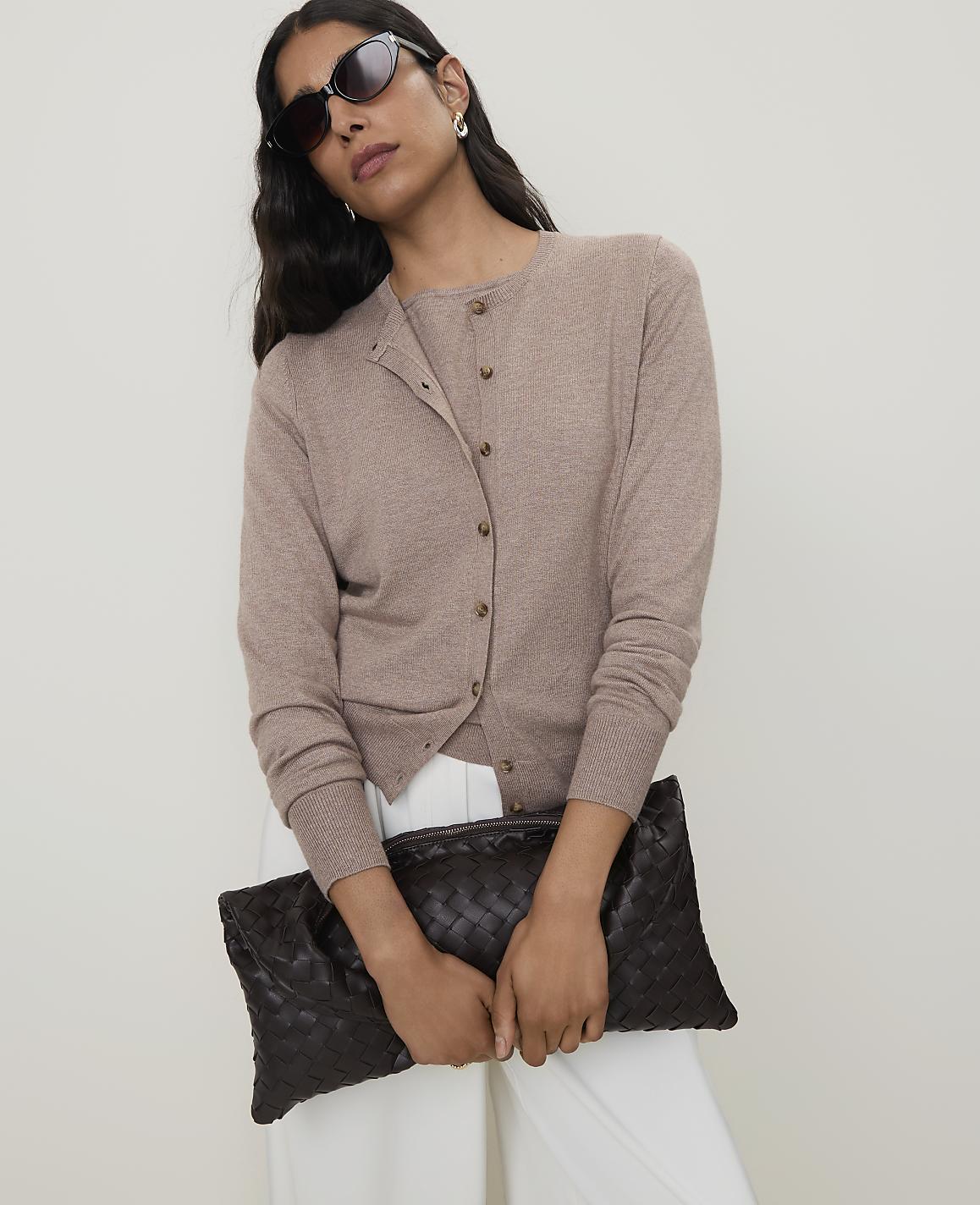 A woman in sunglasses and a beige cardigan holds a dark brown woven clutch in front of a plain white background.