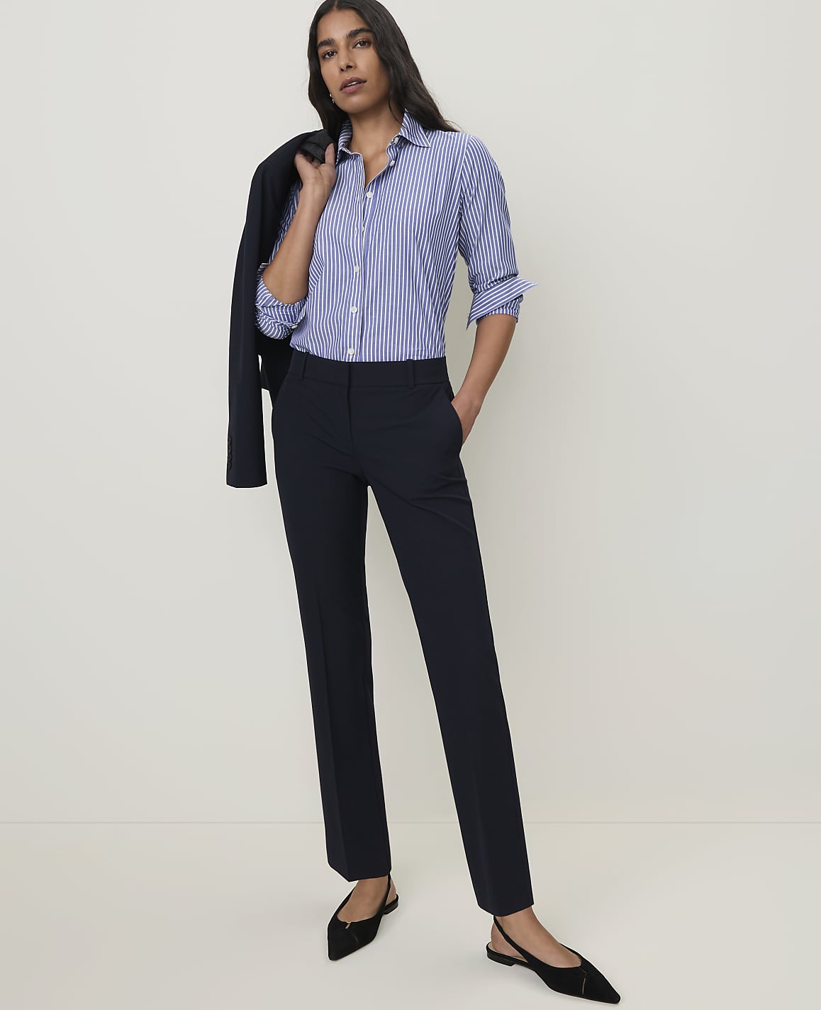 A woman in a striped shirt and dark pants stands against a plain, light background with a dark jacket draped over her shoulder.