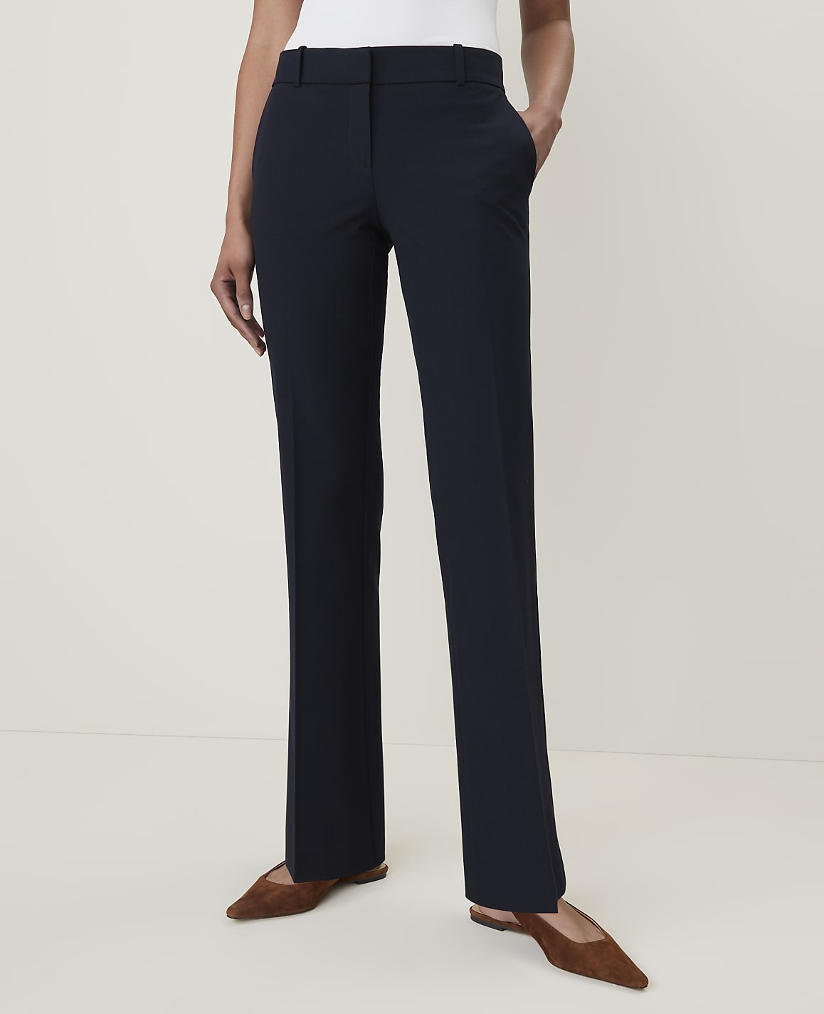 A person stands in dark blue dress pants and brown flats against a plain, light-colored background.