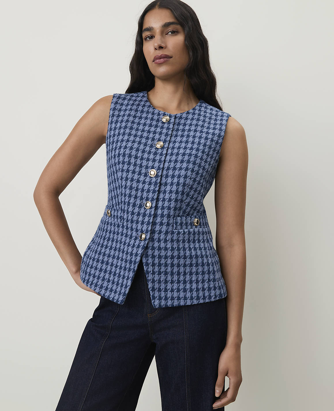 A woman in a blue houndstooth vest and dark jeans stands against a plain, light-colored background.