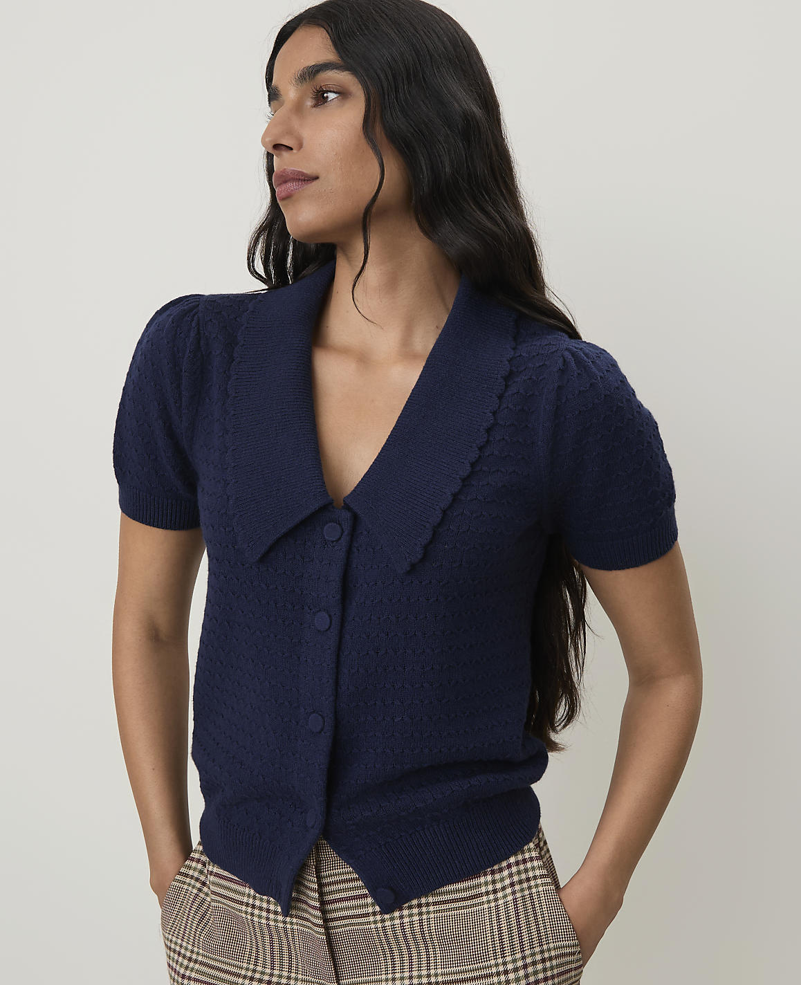 A woman in a navy blue cardigan with puffed sleeves and a scalloped collar stands against a plain, light-colored background, her hands in the pockets of her plaid pants.