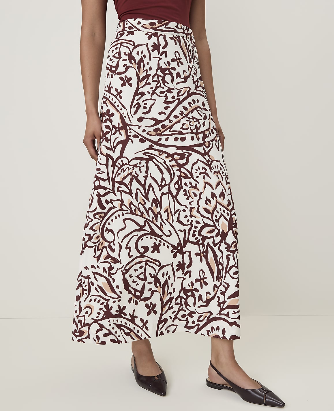 A woman in a patterned skirt and dark flats stands against a plain white background.