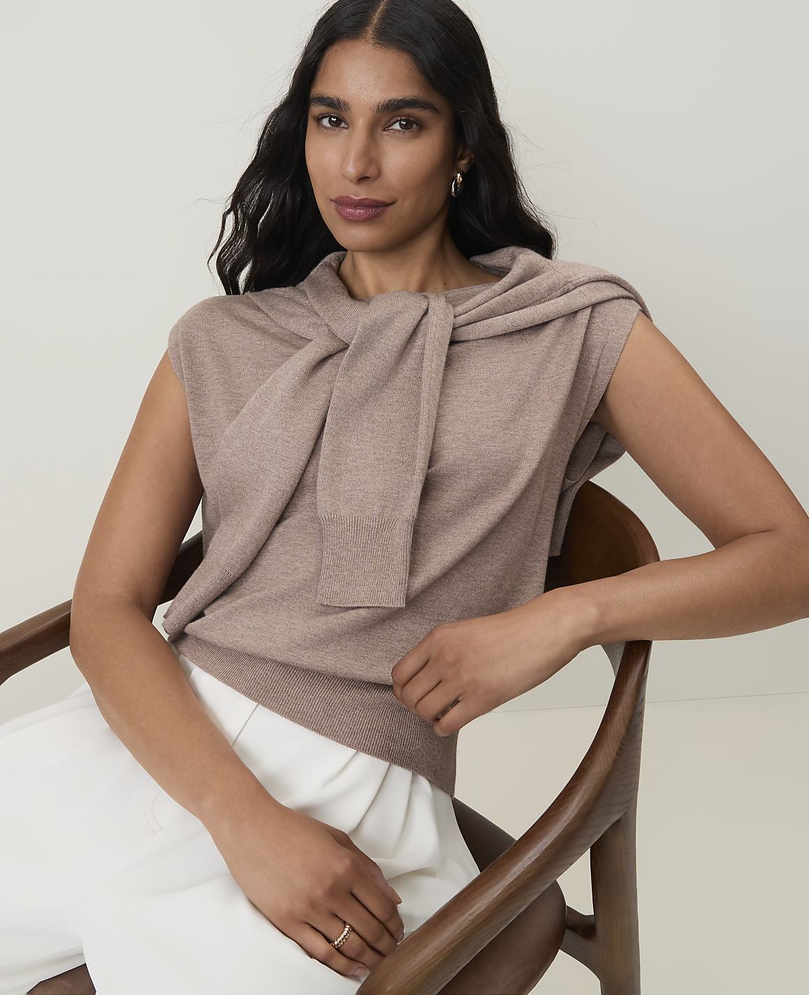 A woman in a taupe sweater and white pants sits in a wooden chair against a plain, light background.