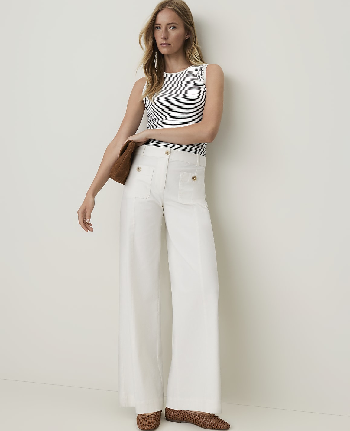 A woman in a striped top and white wide-leg pants stands against a plain white background, holding a brown clutch.