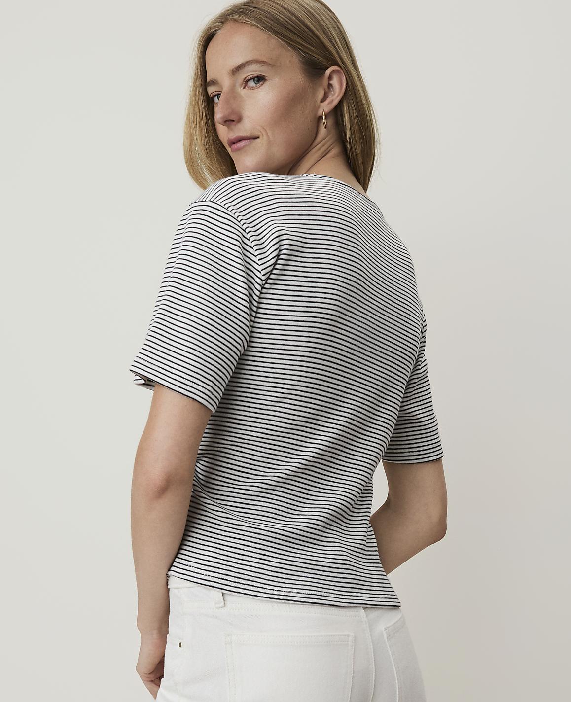 A woman in a striped shirt and white pants stands against a plain, light-colored background.