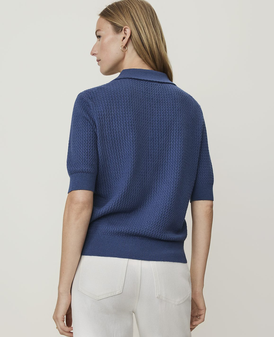 A woman in a blue textured polo shirt and white pants stands against a plain white background.