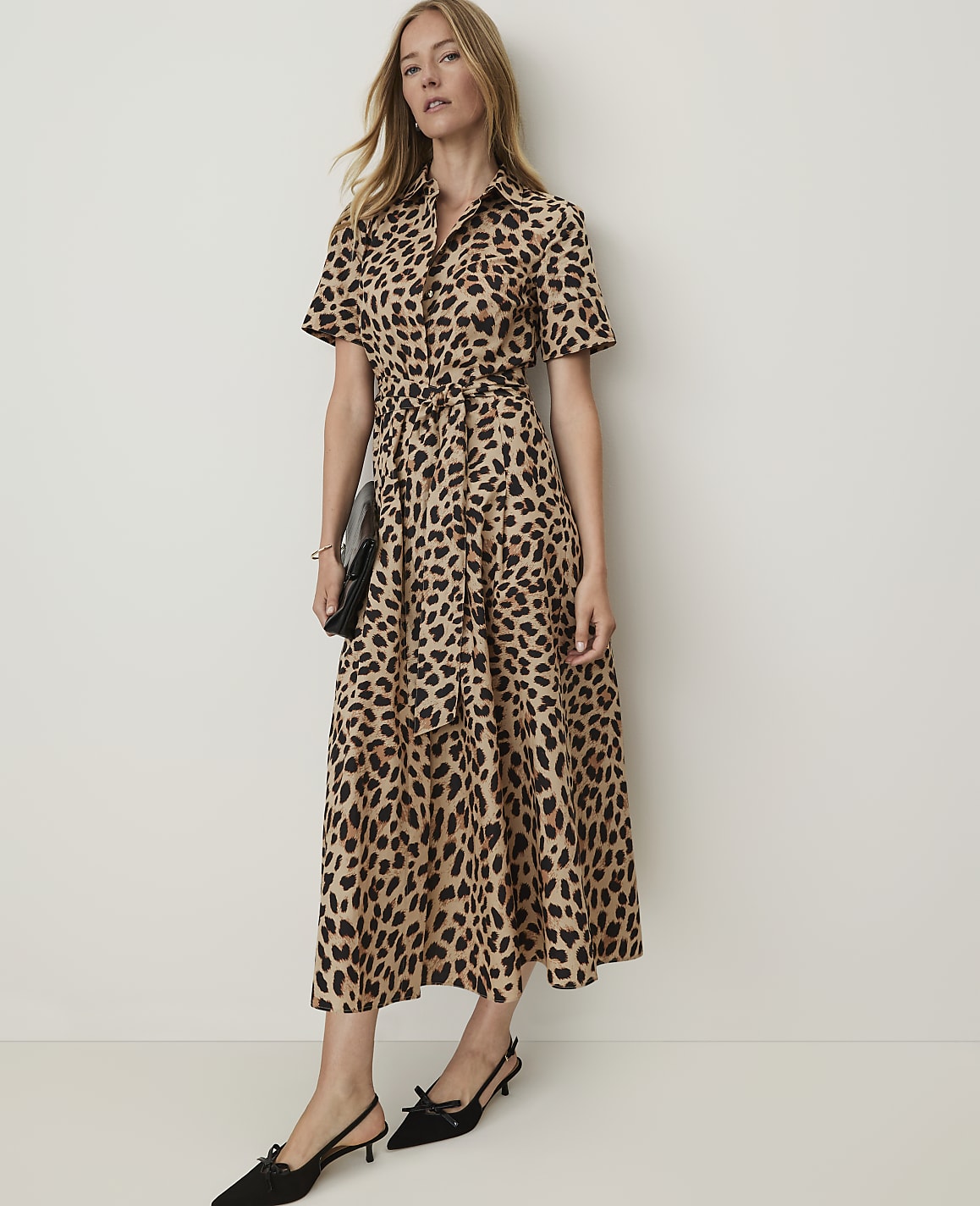 A woman in a leopard-print dress and black kitten heels stands against a plain, light-colored wall, holding a black clutch.