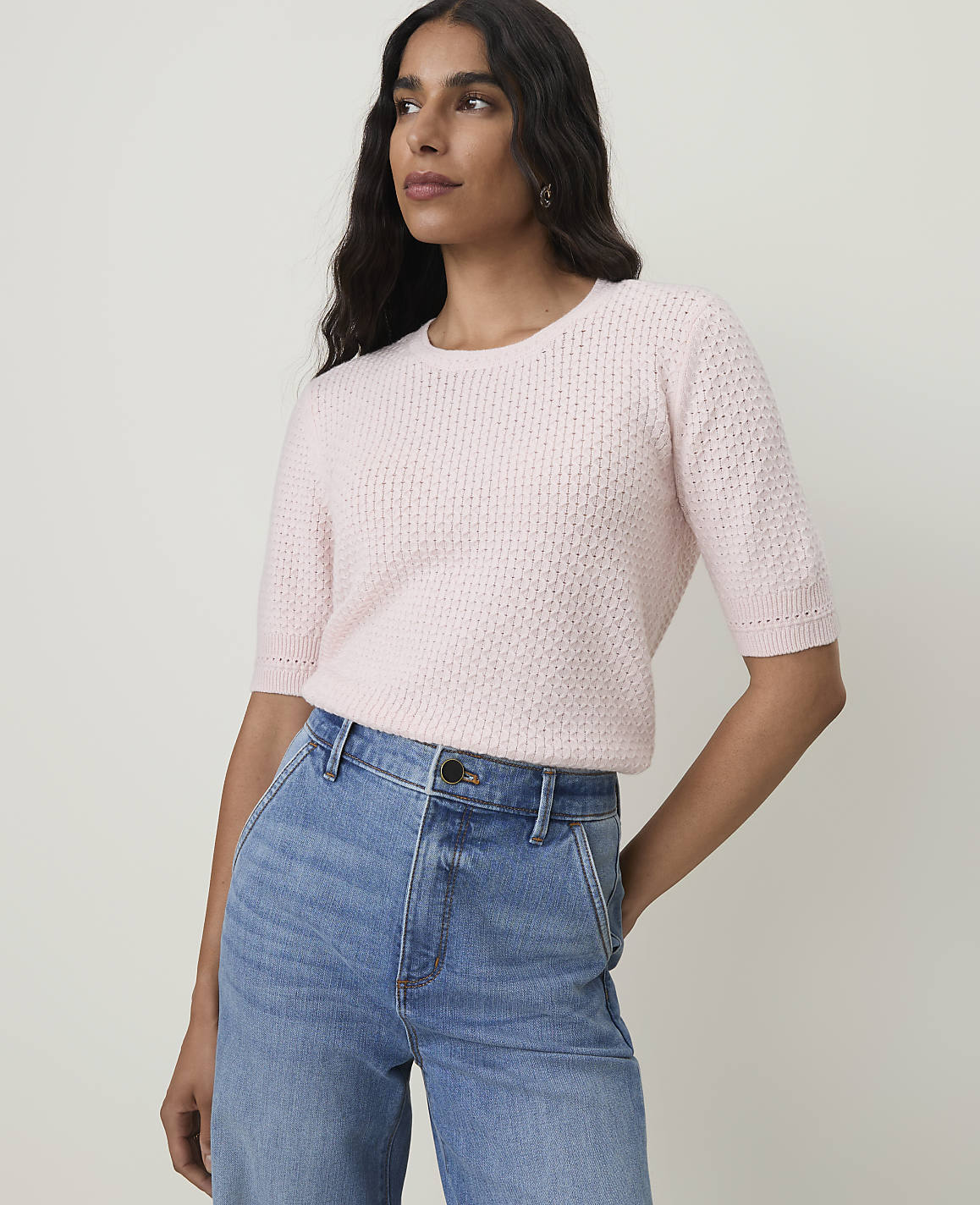 A woman in a pink sweater and blue jeans stands against a plain background.