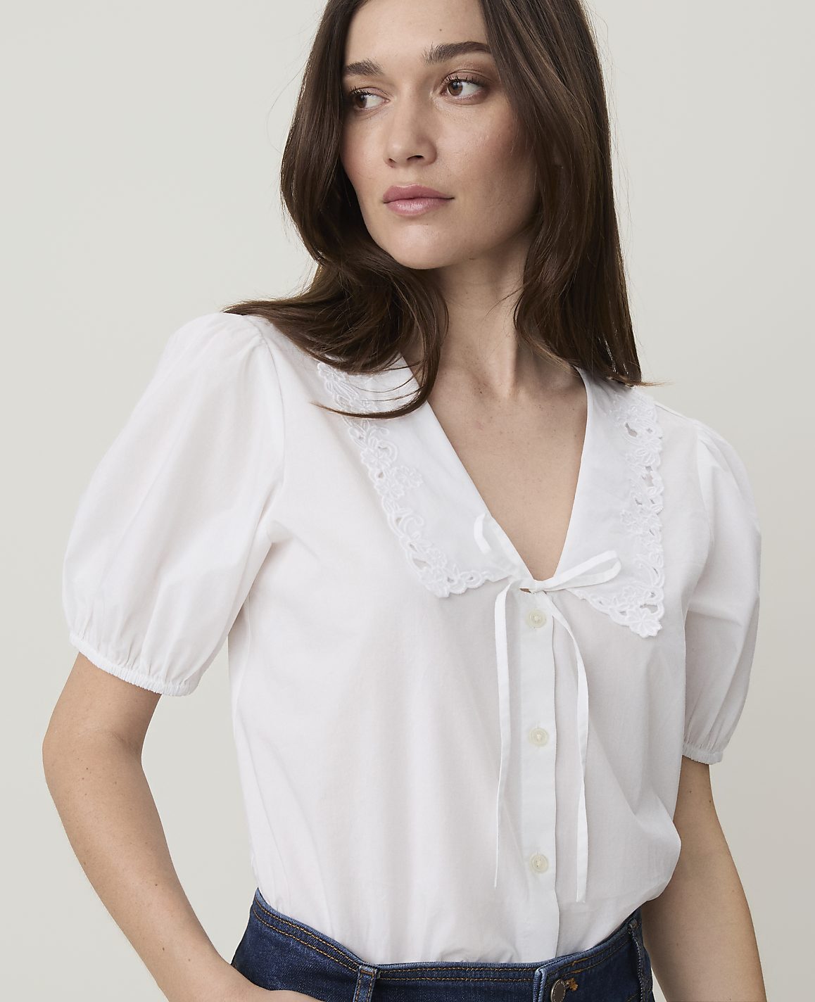 A woman in a white puff-sleeved blouse with a lace collar and a white ribbon tie looks to the side against a plain white background.