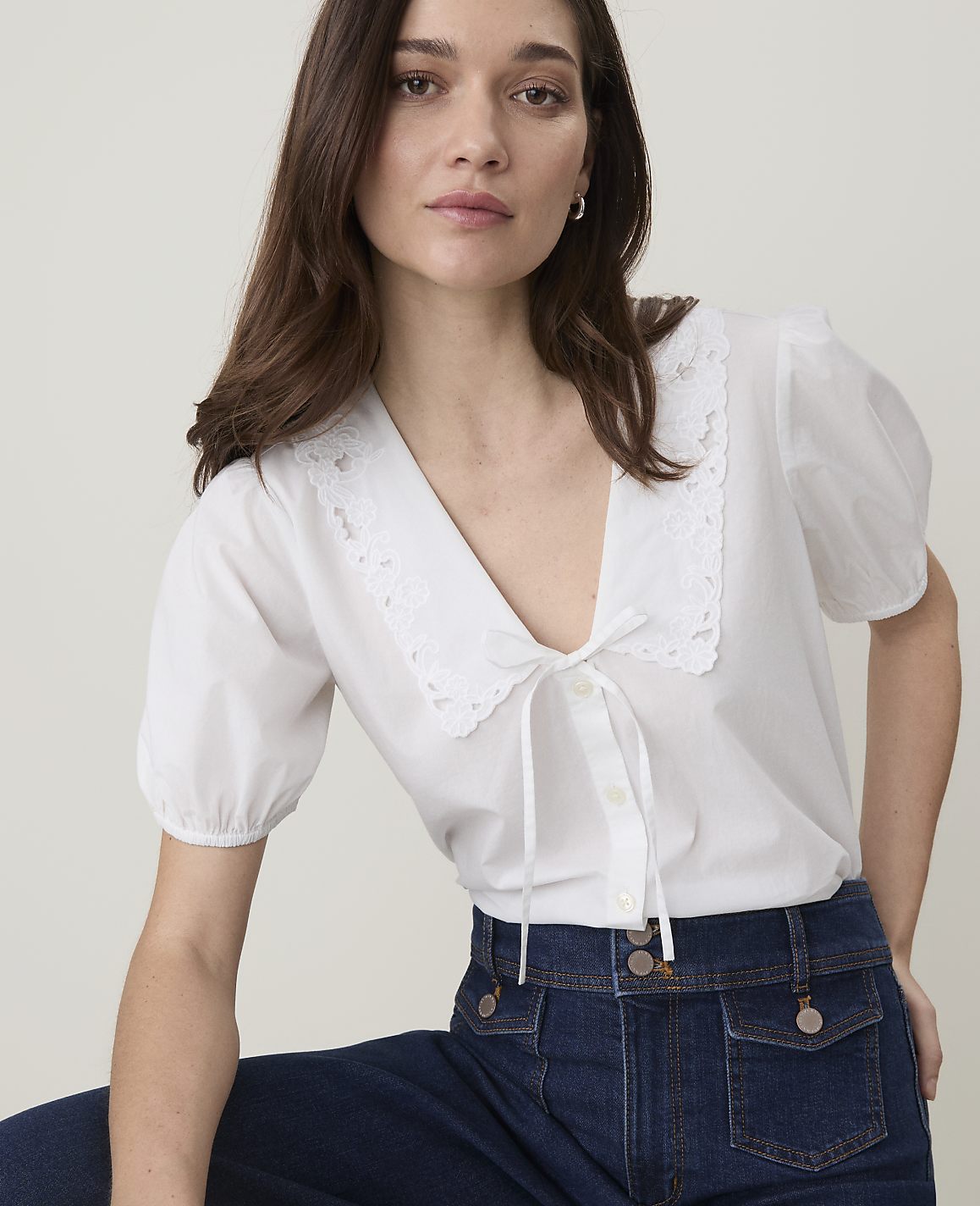 A woman in a white lace-collared blouse and dark jeans poses against a plain, light background.