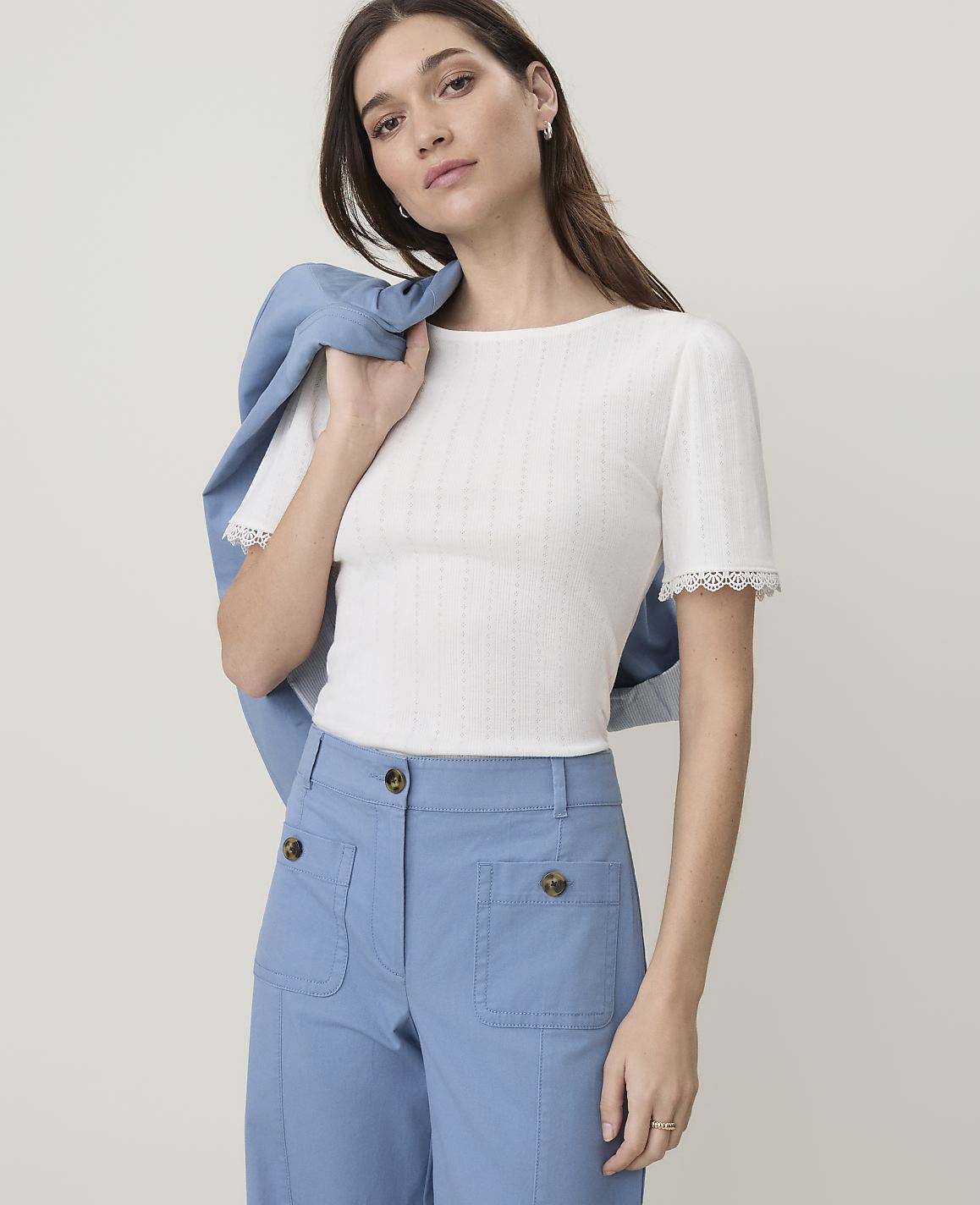 A woman in a white ribbed top and blue pants poses with a blue jacket draped over her shoulder against a plain background.