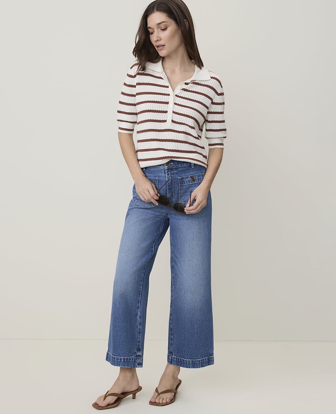 A woman in a striped polo shirt and wide-leg jeans stands against a plain, light-colored background.