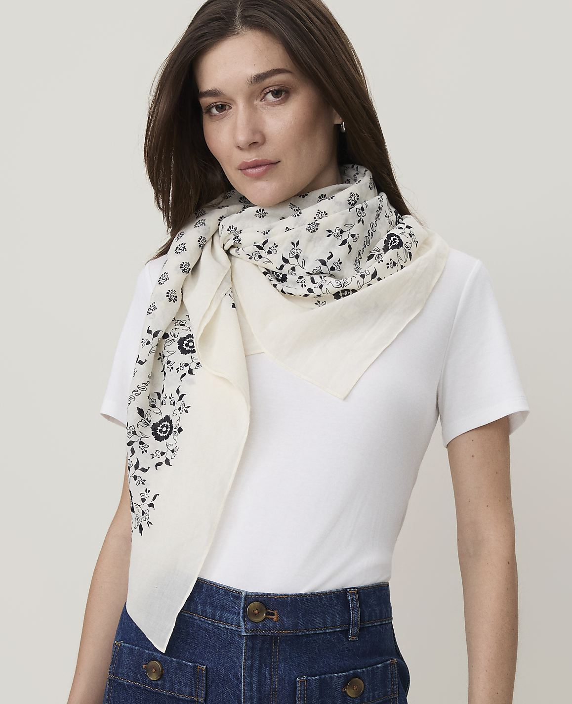 A woman in a white t-shirt and denim skirt models a cream-colored scarf with a black floral pattern against a plain white background.