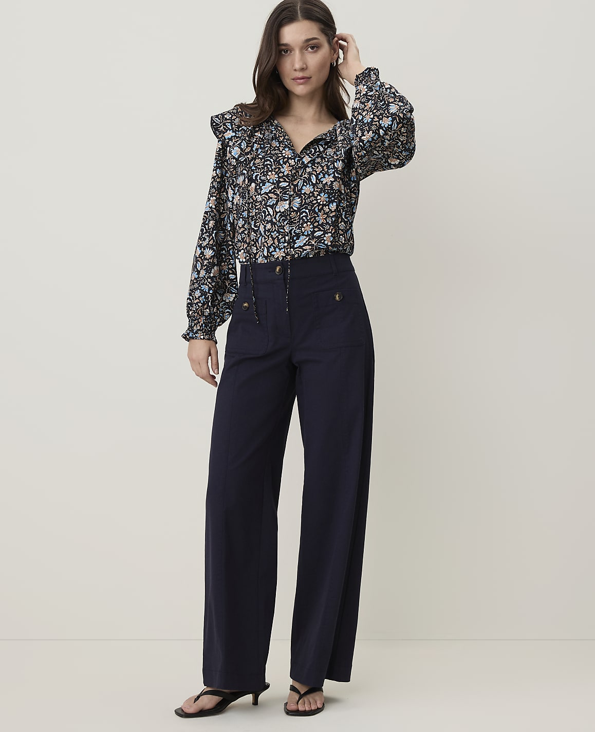 A woman in a floral blouse and navy wide-leg pants stands against a plain, light-colored background.