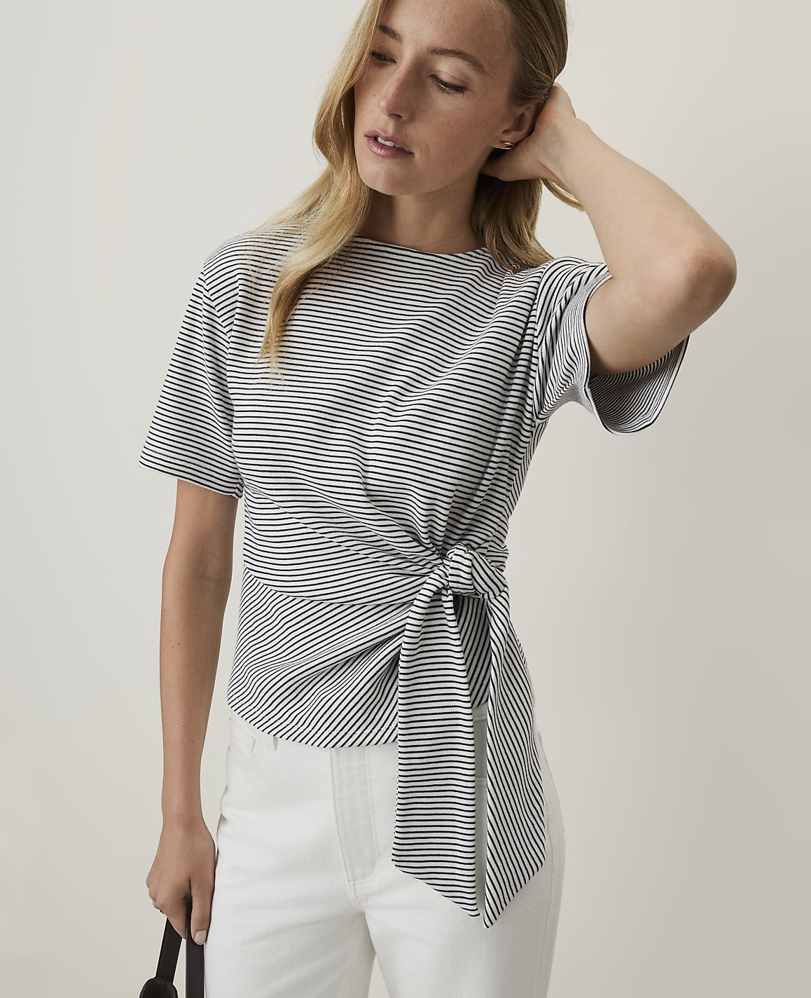 A woman in a striped tie-waist top and white pants stands against a plain, light background.