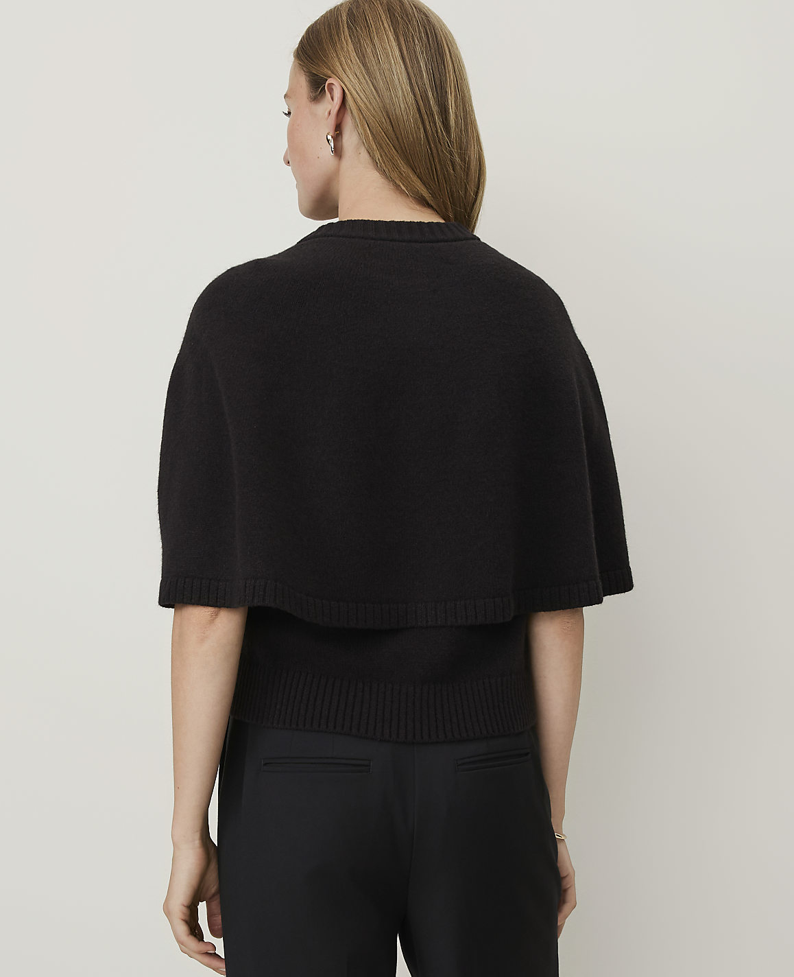 A woman in a black cape sweater and black pants stands against a plain white background.
