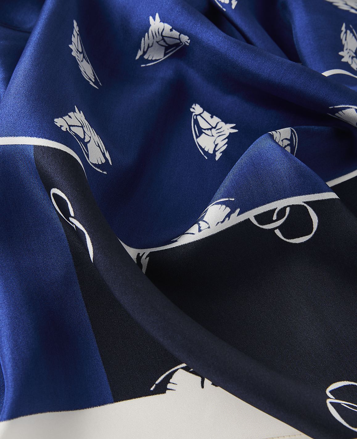A close-up of a blue silk scarf with a pattern of white horse heads and equestrian tack in the foreground, set against a background of darker blue and white sections of the fabric.