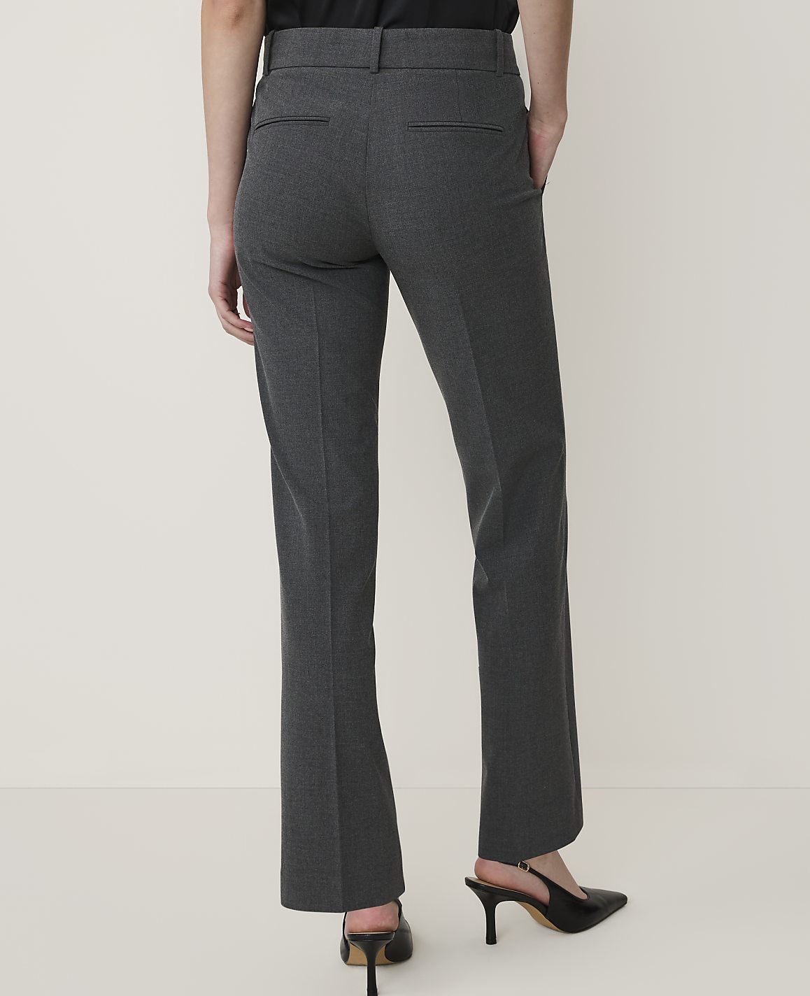 A person wearing dark gray dress pants and black heels stands against a plain, light-colored background.