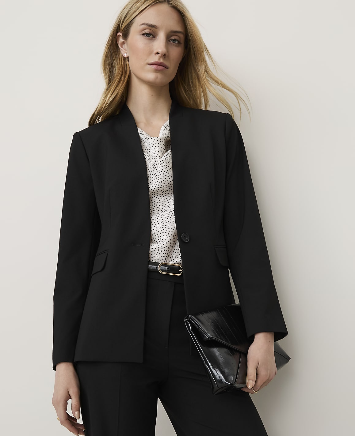 A woman in a black blazer and polka dot blouse holds a black clutch in front of a plain background.