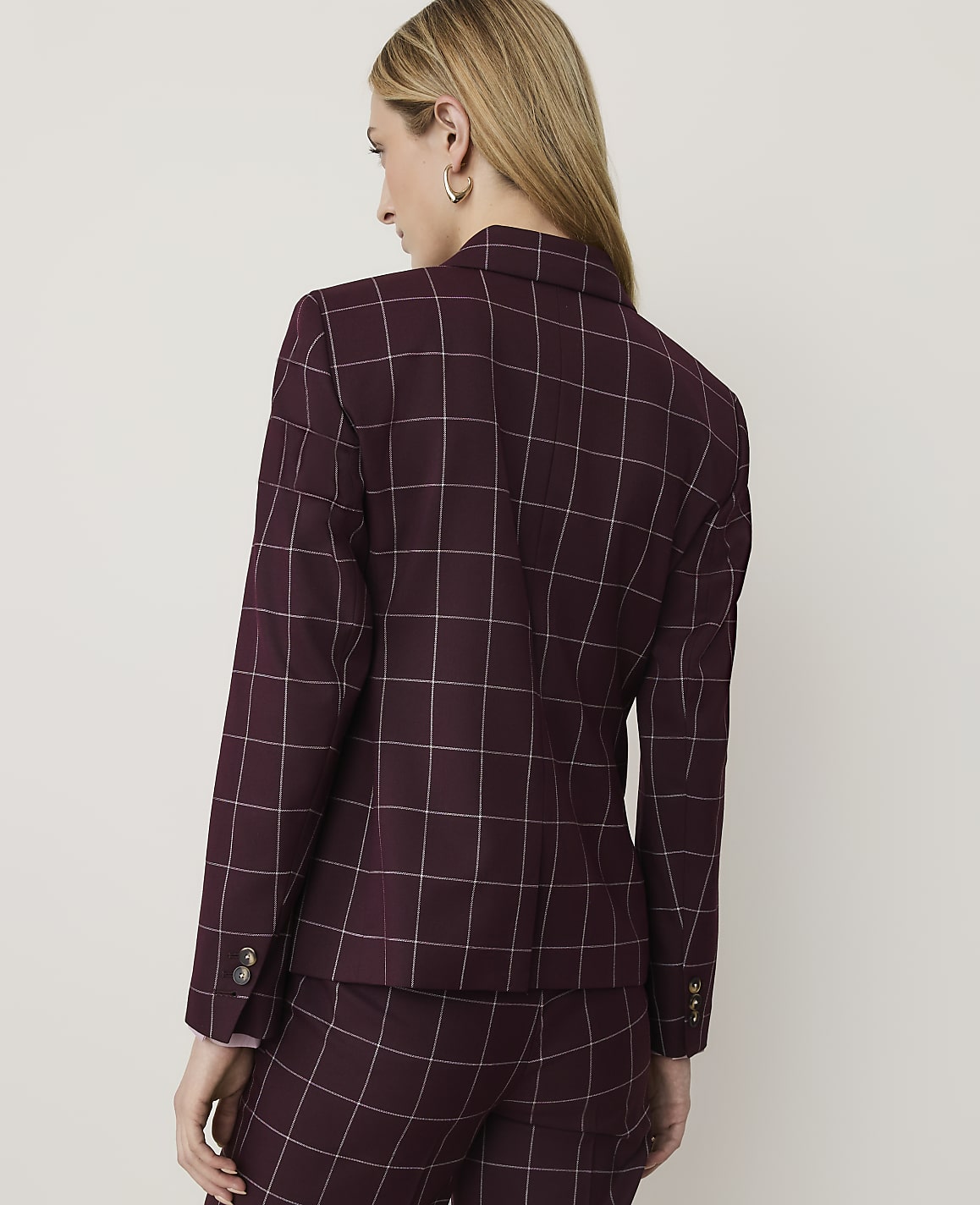 A woman in a burgundy plaid blazer and matching pants stands against a plain, light-colored background.