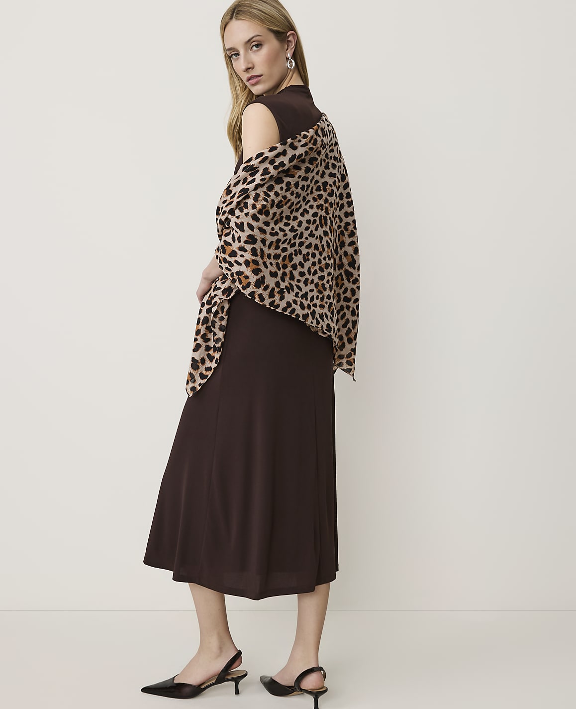 A woman in a brown dress and leopard print shawl stands against a plain white background.