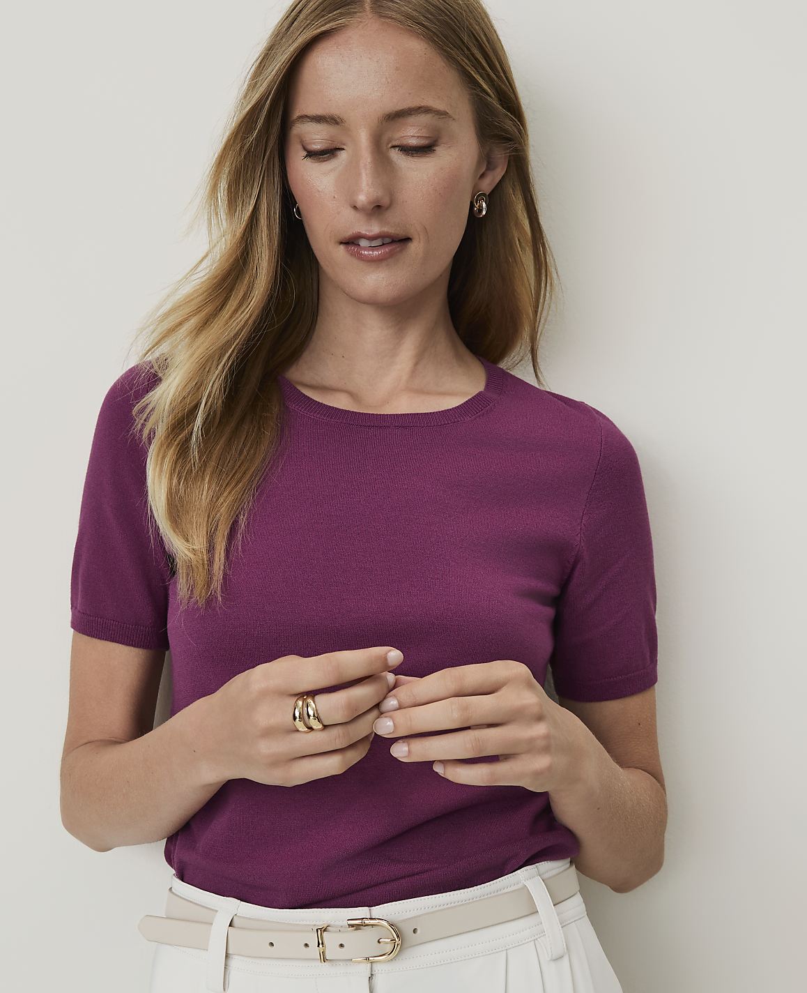A woman in a purple top and white skirt stands against a plain background.