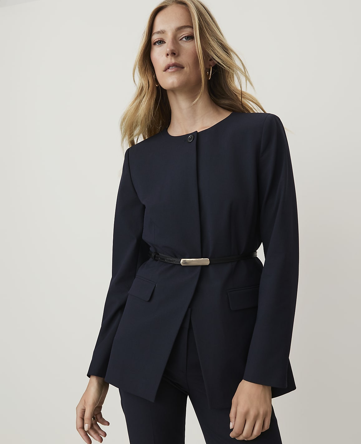 A woman in a navy blazer cinched with a belt stands against a plain, light background.