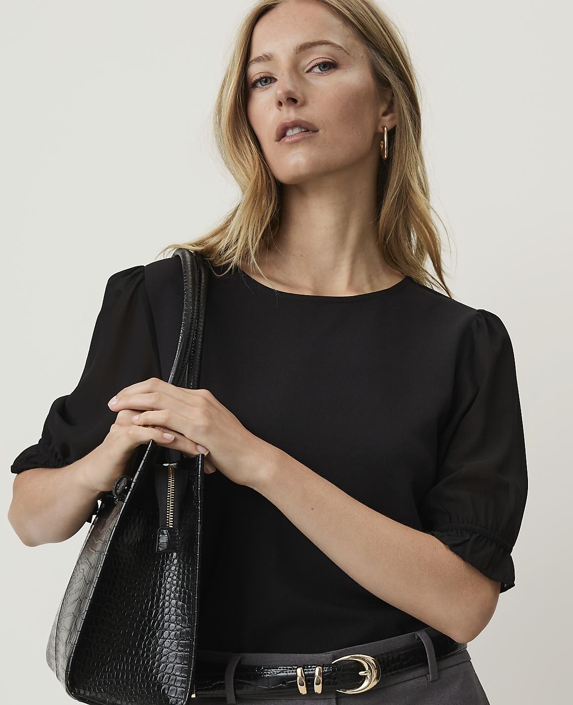 A woman in a black top and grey pants holds a black textured handbag against a plain background.