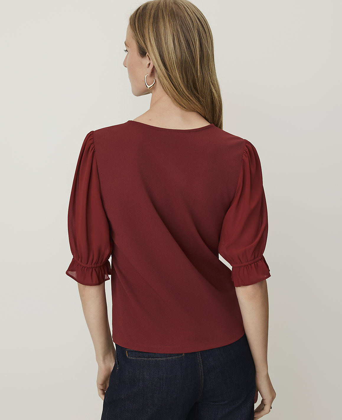 A woman in a maroon top with puffed sleeves and dark jeans stands against a plain white background.