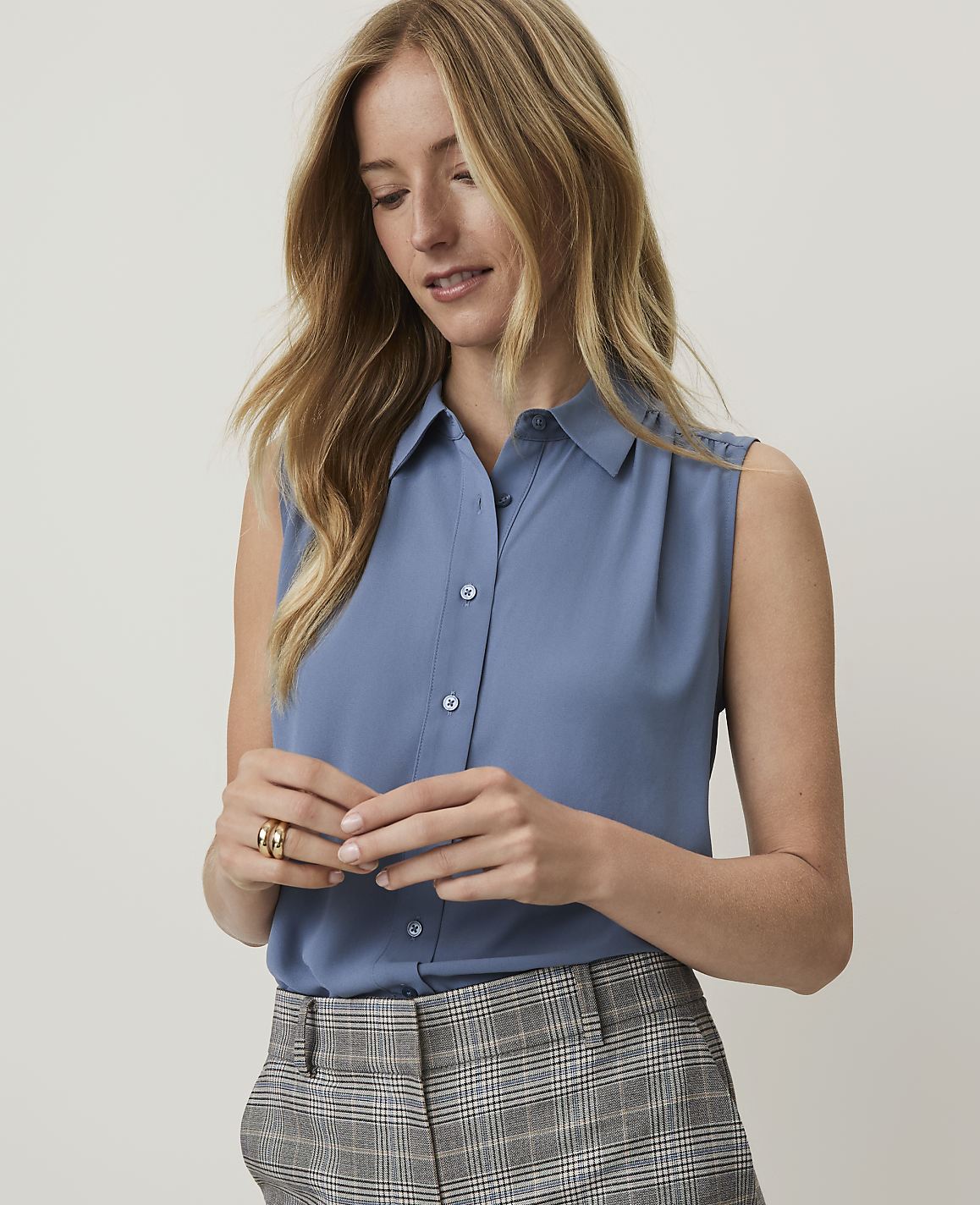 A woman in a blue sleeveless blouse and plaid pants looks down at her hands in the foreground, with a plain background behind her.