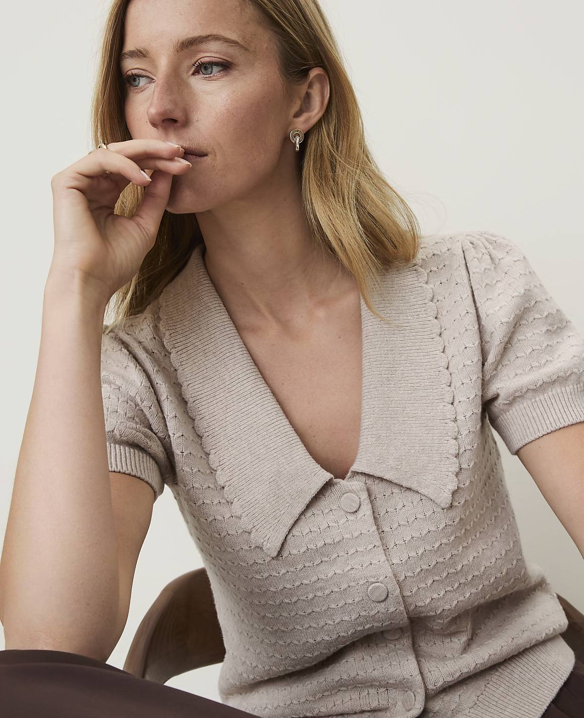 A woman in a beige collared sweater with textured details rests her chin on her hand, with a blurred brown background.