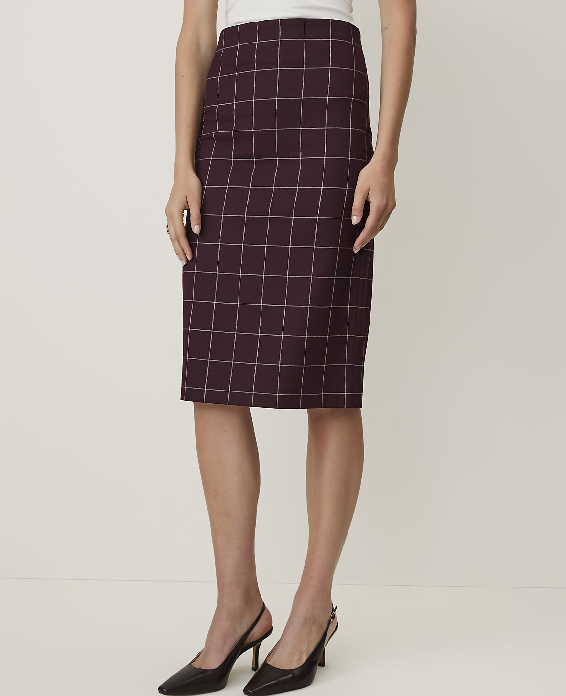 A woman in a burgundy plaid pencil skirt and black slingback heels stands against a plain white background.
