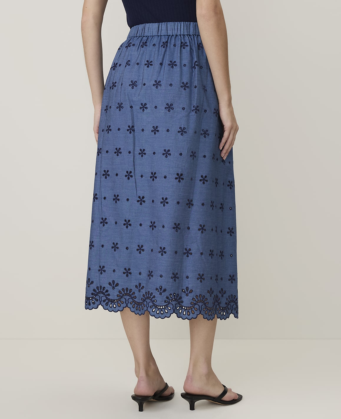 A woman models a blue eyelet midi skirt with a floral pattern and scalloped hem against a plain background.
