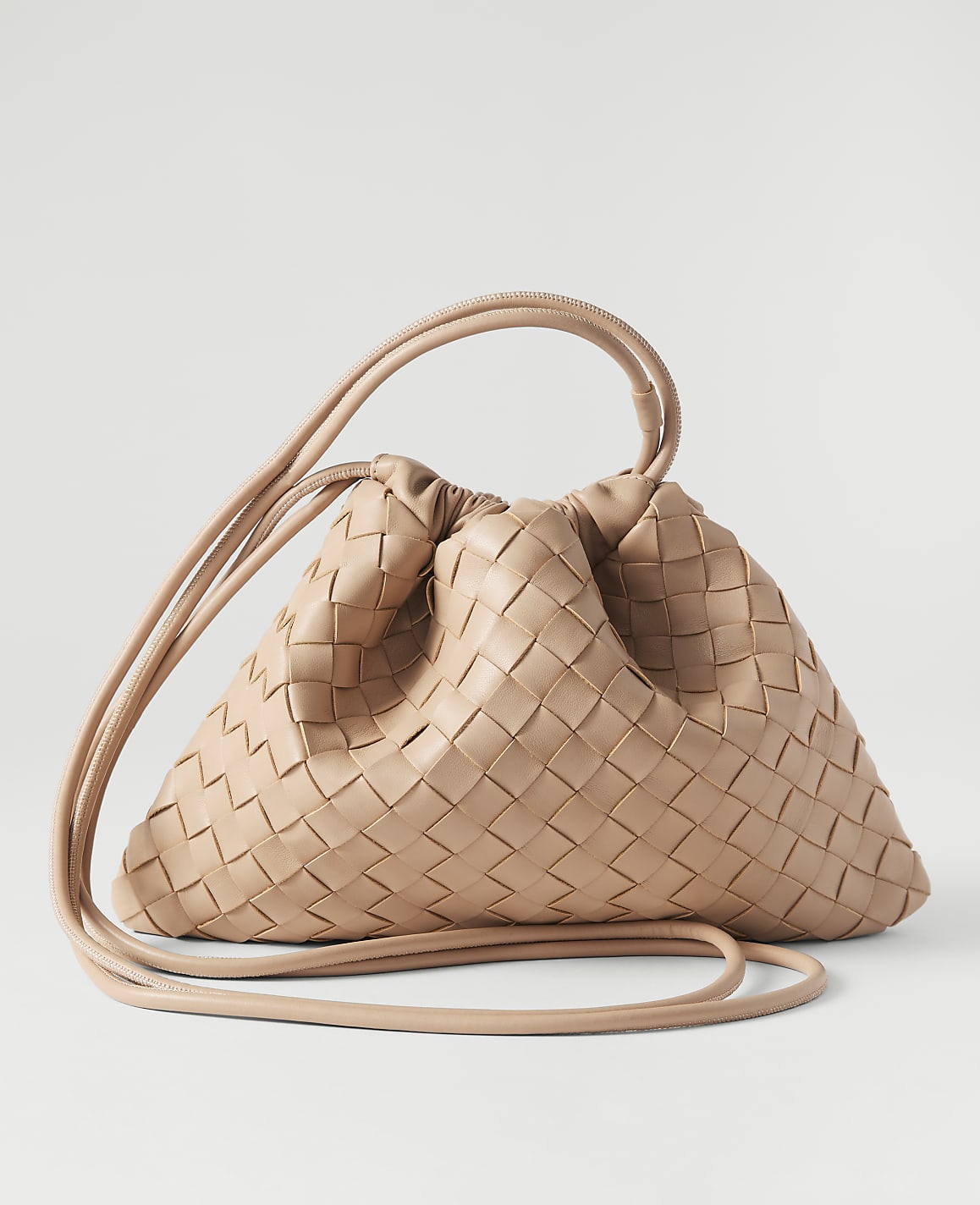 A light beige woven leather handbag with a drawstring closure and long strap rests on a plain white background.