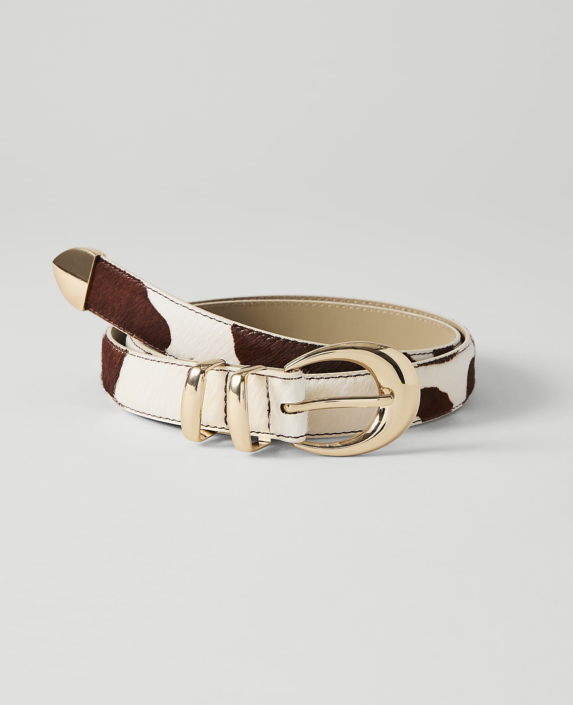 A cow-print belt with gold hardware lies on a plain white background.
