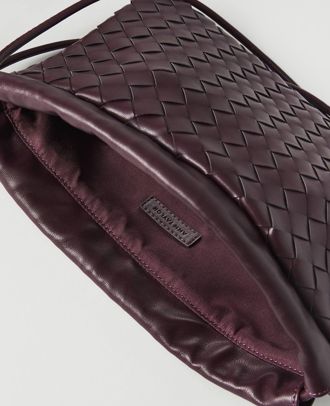 A woven burgundy leather clutch with a satin lining and a brand tag is shown against a white background.