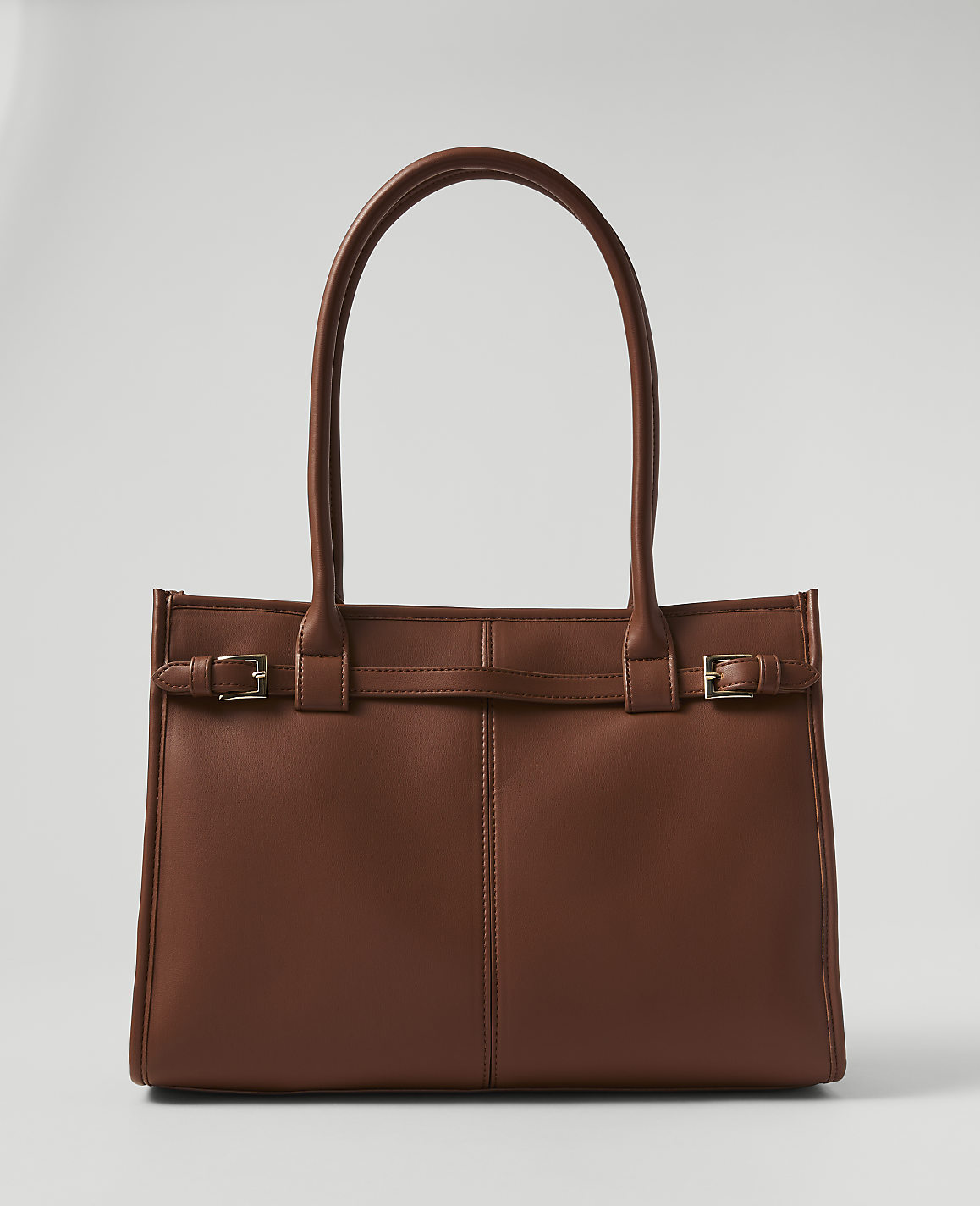 A brown leather tote bag with buckle details sits in the foreground against a plain, light gray background.