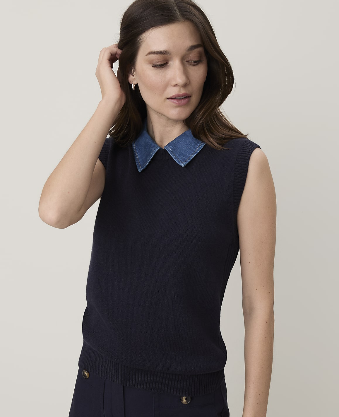 A woman in a navy sweater vest over a denim collar stands against a plain white background.