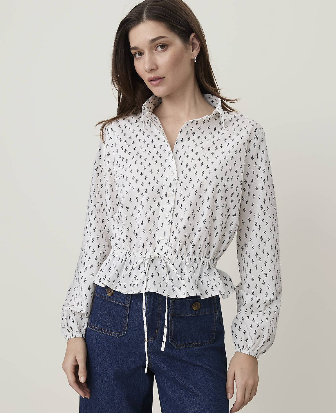A woman in a white patterned blouse and dark blue pants stands against a plain background.